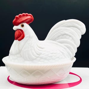 May include: A white milk glass hen on a nest dish featuring a rooster lid with a bright red comb and wattle. The base is designed with a textured basket-weave pattern. The rooster has a detailed molded feather design and a prominent tail.
