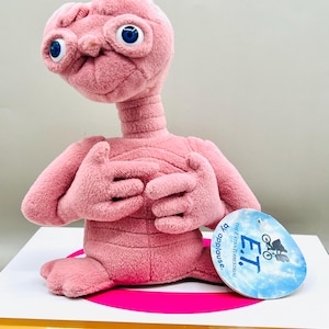 May include: A pink plush E.T. doll with large blue eyes and a long neck. The doll has outstretched arms and is seated. A tag with the text "E.T. The Extra Terrestrial" is attached.