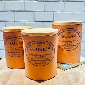 May include: Three orange ceramic canisters with wooden lids. The canisters are labeled "Flour", "Cookies", and "Sugar" in black text. Each canister has the text "The Original Suffolk Canister" and "Henry Watson Pottery Founded 1800".