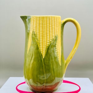 May include: A ceramic pitcher with a corn on the cob design. The pitcher features a yellow handle and spout, with a green and yellow corn design on the body. The base is brown. The pitcher is likely used for serving beverages.