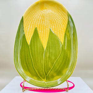 May include: A ceramic serving platter shaped like an ear of corn. The platter features a yellow corn cob design at the top and green leaves at the bottom. The platter is oval-shaped and rests on a gold-colored metal stand with a pink ribbon.