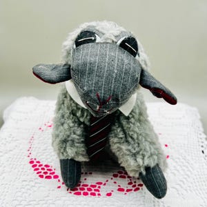 May include: A plush sheep toy featuring a grey pinstriped fabric face and limbs, a fluffy grey body, a white collar, and a red and black striped necktie. The sheep has dark fabric ears with red lining and stitched facial features.