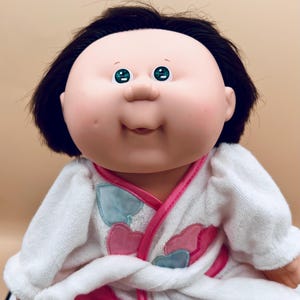 May include: A Cabbage Patch Kids doll with black hair and green eyes, wearing a white terry cloth robe with pink and blue heart-shaped appliqués. The robe has pink trim and a pink heart-shaped patch with the letters "CP" on it.
