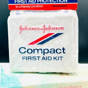 May include: A white Johnson & Johnson Compact First Aid Kit in a clear plastic wrapper. The kit has red and blue accents and the text "FIRST AID PROTECTION In a Handy Location". The kit is on top of a book.