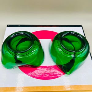 Puede incluir: Dos cuencos decorativos de vidrio verde con una forma &uacute;nica. Los cuencos tienen un acabado brillante y un patr&oacute;n de remolino verde oscuro en el interior. Los cuencos est&aacute;n sobre una superficie blanca con un c&iacute;rculo rojo.