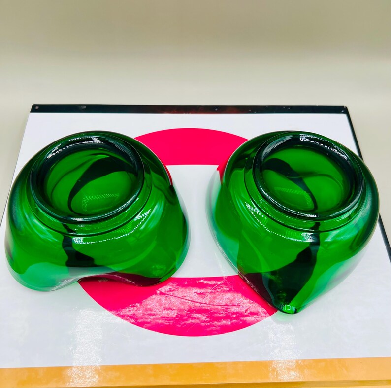 Puede incluir: Dos cuencos decorativos de vidrio verde con una forma &uacute;nica. Los cuencos tienen un acabado brillante y un patr&oacute;n de remolino verde oscuro en el interior. Los cuencos est&aacute;n sobre una superficie blanca con un c&iacute;rculo rojo.