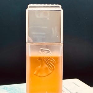 May include: A rectangular perfume bottle with a clear, frosted design. The bottle features a raised profile of a woman's face and hair. The liquid inside is a warm orange color. The top is a clear, rectangular cap with a silver border.
