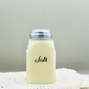 Può includere: Saliera vintage color crema con coperchio in metallo argentato. La parola "Salt" è scritta in corsivo sulla parte anteriore della saliera. La saliera è rettangolare con linee verticali ed è appoggiata su una tovaglietta di pizzo bianco.