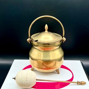 May include: A vintage brass sugar bowl with a lid and handle, on a book. The bowl has a small spoon and sits on three small legs. A light-coloured, round object with a metal handle is next to the bowl.