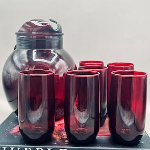 May include: A set of six ruby red drinking glasses and a matching pitcher with a lid. The glasses are tall and cylindrical. The pitcher has a rounded body and a curved handle. The items are displayed on a book.