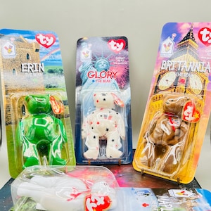 Puede incluir: Tres peluches coleccionables Ty Beanie Baby en su embalaje original. Los osos se llaman Erin, Glory y Britannia. El oso Erin es verde, Glory es blanco con estrellas rojas y azules, y Britannia es marrón. También se ve un peluche blanco.