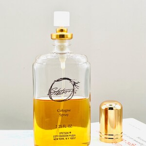 May include: A clear glass cologne bottle with a gold cap and a white top. The bottle contains a golden-colored liquid and has the brand name "Stetson" printed on the front. The bottle is labeled "Cologne Spray 2.25 FL. OZ."