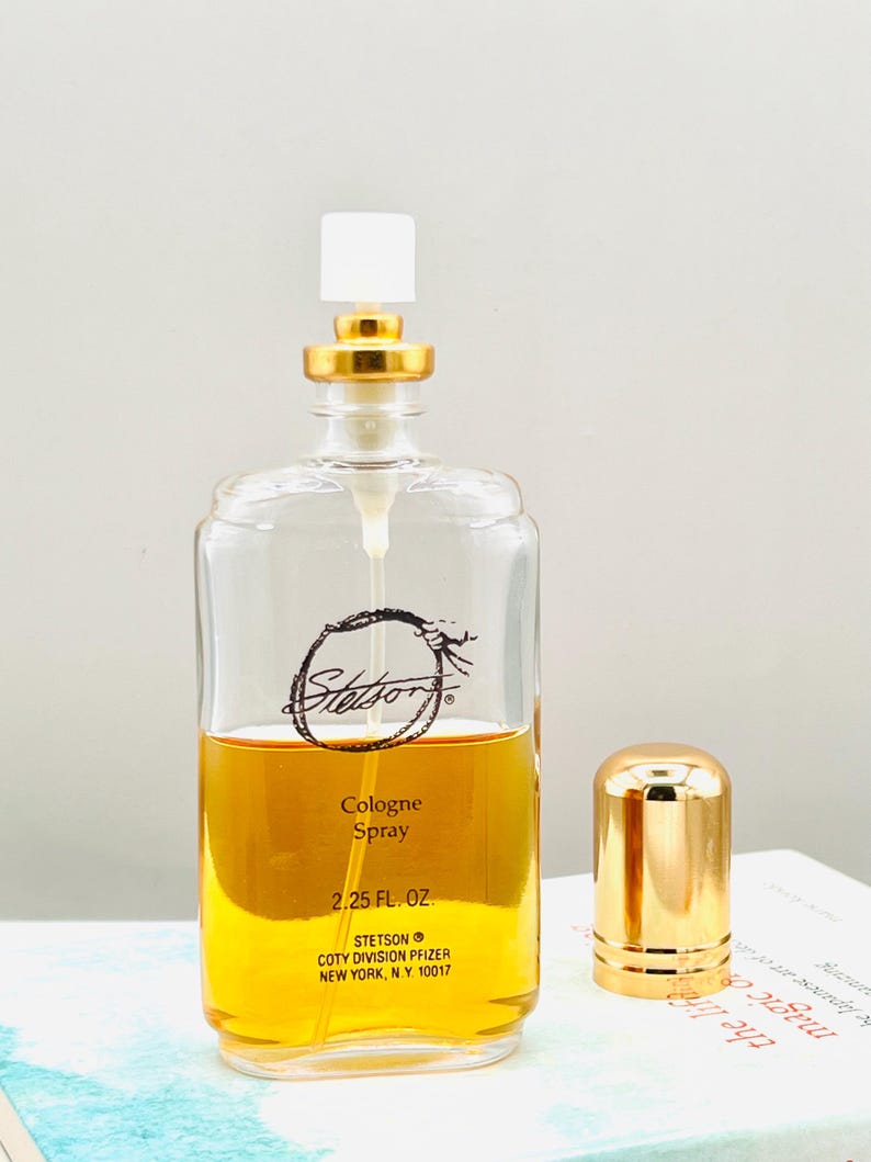May include: A clear glass cologne bottle with a gold cap and a white top. The bottle contains a golden-colored liquid and has the brand name "Stetson" printed on the front. The bottle is labeled "Cologne Spray 2.25 FL. OZ."