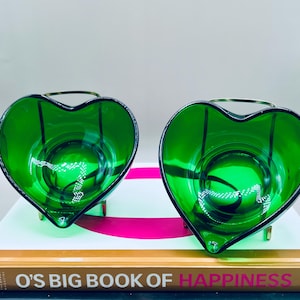 Puede incluir: Dos candelabros de vidrio verde esmeralda en forma de coraz&oacute;n. Cada soporte tiene un asa y una base de metal dorado. Los candelabros est&aacute;n sobre un libro titulado "O's Big Book of Happiness".