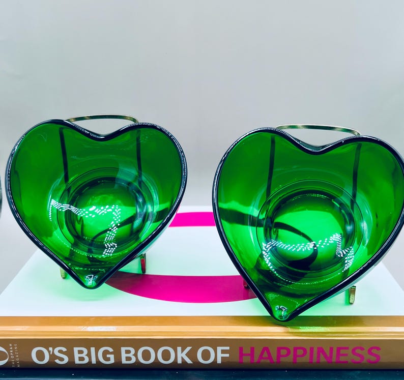 Puede incluir: Dos candelabros de vidrio verde esmeralda en forma de coraz&oacute;n. Cada soporte tiene un asa y una base de metal dorado. Los candelabros est&aacute;n sobre un libro titulado "O's Big Book of Happiness".
