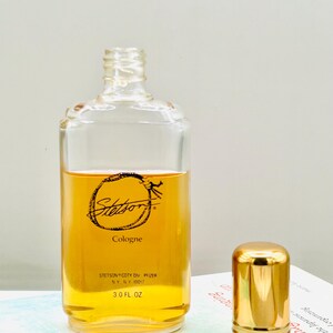 May include: A clear glass bottle of Stetson cologne with a gold cap. The bottle contains a golden-colored liquid and has the Stetson logo and text printed on the front. The bottle is 3.0 fl oz.