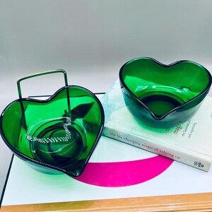 Puede incluir: Dos cuencos de vidrio verde esmeralda en forma de coraz&oacute;n. Un cuenco tiene un asa de metal y contiene un collar plateado. El otro cuenco est&aacute; vac&iacute;o. Ambos cuencos est&aacute;n sobre una superficie blanca con un libro.