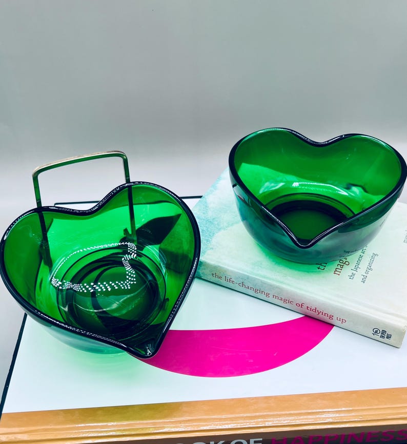 Puede incluir: Dos cuencos de vidrio verde esmeralda en forma de coraz&oacute;n. Un cuenco tiene un asa de metal y contiene un collar plateado. El otro cuenco est&aacute; vac&iacute;o. Ambos cuencos est&aacute;n sobre una superficie blanca con un libro.