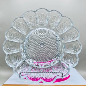 May include: Clear glass serving plate with a scalloped edge and a textured, dotted center. The plate is supported by a metal stand and rests on a pink surface. The plate's design resembles a flower.
