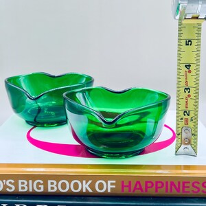 Puede incluir: Dos cuencos de vidrio verde esmeralda con borde en forma de coraz&oacute;n. Los cuencos miden aproximadamente 5 cm de altura. Una cinta m&eacute;trica amarilla y negra est&aacute; en el fondo.