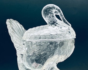 May include: Clear glass turkey-shaped covered dish. The dish features detailed feather and body patterns. The turkey has a curved neck and a lid that fits over the body. The dish is sitting on a book.