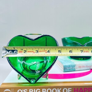 Puede incluir: Un plato de vidrio verde en forma de coraz&oacute;n con un asa de metal dorado, que mide aproximadamente 10 cm de ancho. Un segundo plato de vidrio verde es visible en el fondo. Los platos est&aacute;n sobre libros, incluido "O's Big Book of Happiness."