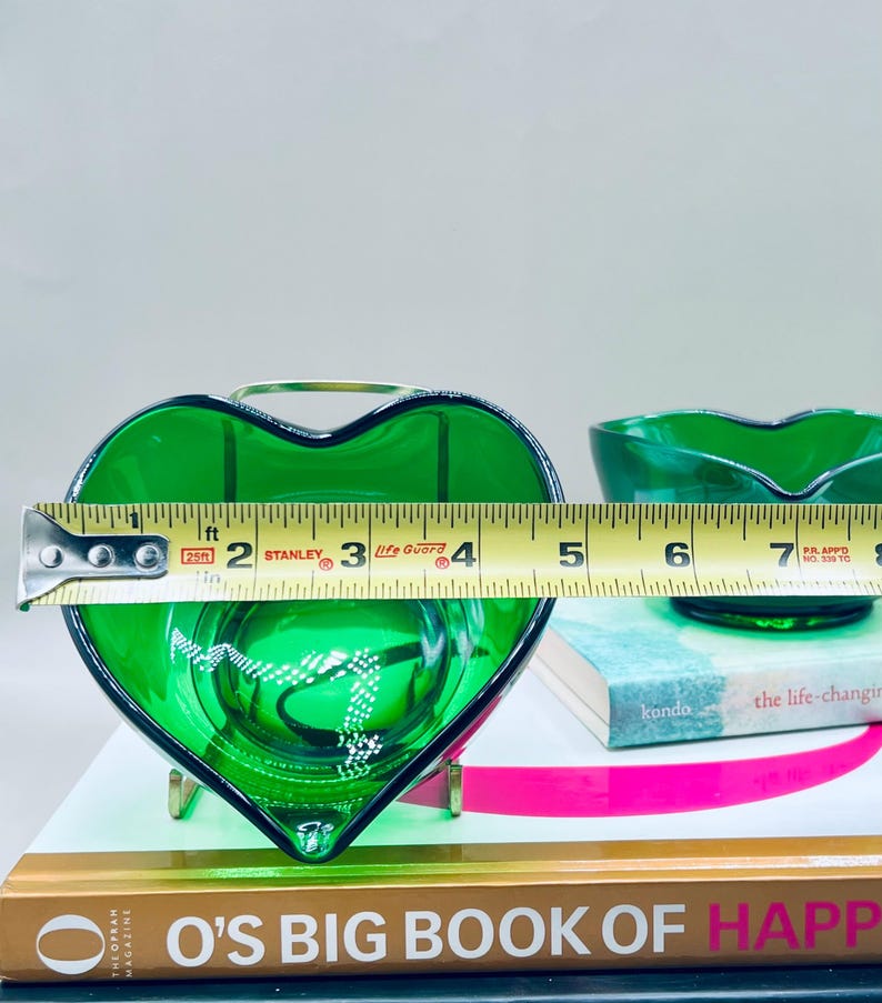 Puede incluir: Un plato de vidrio verde en forma de coraz&oacute;n con un asa de metal dorado, que mide aproximadamente 10 cm de ancho. Un segundo plato de vidrio verde es visible en el fondo. Los platos est&aacute;n sobre libros, incluido "O's Big Book of Happiness."