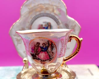 May include: A miniature porcelain teacup and saucer set with a gold trim and a decorative scene of a couple dancing. The cup and saucer are set on a gold-colored stand against a bright pink background.