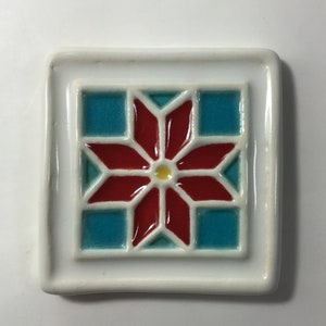 May include: A white ceramic tile with a red and blue geometric design. The design features a starburst pattern with a yellow center.
