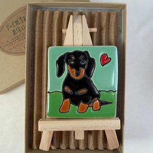 May include: A square ceramic tile featuring a hand-painted black and tan dachshund on a light green background with a small red heart, displayed on a miniature wooden easel inside a cardboard gift box with corrugated paper lining.