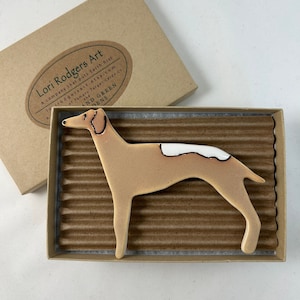 May include: A ceramic dog figurine in a brown box. The dog is tan with a white patch on its back. The box has a label that says "Lori Rodgers Art" and "A company that puts Earth first".