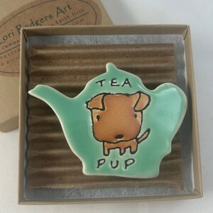 May include: A teal ceramic teapot shaped like a dog with the words "TEA PUP" on it. The dog is brown with black eyes and a black nose.