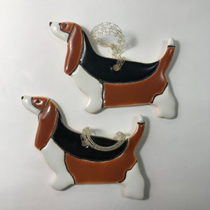 May include: Two ceramic ornaments shaped like basset hounds. The ornaments are painted in brown, black, and white. They have a gold string attached for hanging.