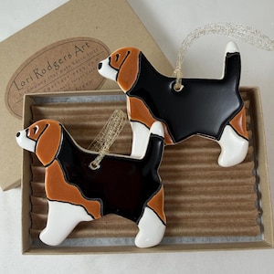 May include: Two ceramic beagle dog ornaments featuring a tri-color pattern of black, brown, and white. Each ornament has a gold hanging loop and is presented in a cardboard box with a label reading Lori Rodgers Art, a company that puts Earth first.