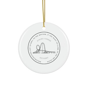 May include: A white ceramic ornament with a gold hanging string. The ornament has a black line drawing of a roller coaster and the text "Rollercoasters Got to Roll to the Bottom, If You Want to Climb to the Top Again. String Cheese Incident."
