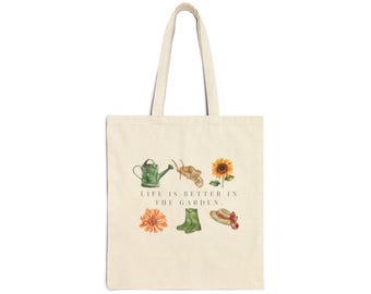 May include: A beige tote bag with a green watering can, a wheelbarrow, a sunflower, a flower, green rain boots, and a straw hat. The text "LIFE IS BETTER IN THE GARDEN." is printed on the bag.