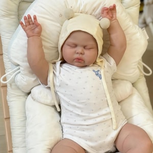 May include: A lifelike baby doll dressed in a cream hat with pom-poms and a white onesie featuring a blue animal design. The doll is lying on a white padded surface with its arms raised. The baby's eyes are closed.