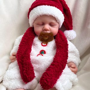 May include: A baby doll wearing a festive Christmas outfit. The doll is dressed in a white plush onesie, a red and white Santa hat, and a red scarf. It has a brown pacifier and Christmas-themed buttons.