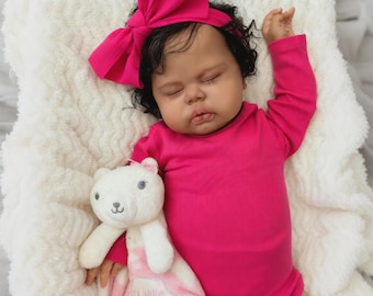 May include: A baby doll dressed in a vibrant pink onesie and a matching bow headband, asleep on a soft, white blanket. The doll clutches a white teddy bear comforter with the words "Snuggle Buddy" embroidered on it.