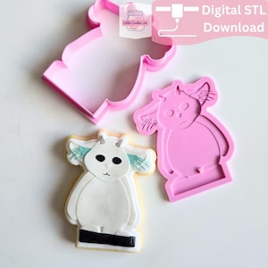 May include: A pink plastic cookie cutter and a matching pink plastic stamp featuring a creature with large ears and small horns, alongside a decorated cookie shaped like the same creature with white icing, black eyes, and teal ear accents.