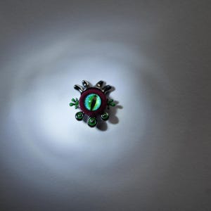 May include: A small, silver-colored pin with a red and green eye design. The eye has a green iris and a black pupil. The pin has four small green leaves around the eye.