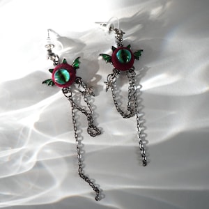 May include: A pair of silver dangle earrings with red and green eye-shaped charms and silver chains.