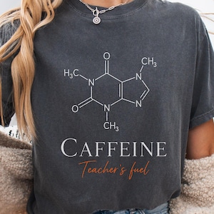 May include: A charcoal gray short-sleeve t-shirt featuring a white chemical structure diagram of a caffeine molecule. Below the diagram, the word CAFFEINE is printed in white capital letters, followed by the text Teacher's fuel in orange script.