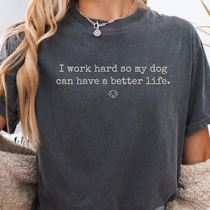 Puede incluir: Una camiseta gris carbón de manga corta con el texto impreso en blanco "I work hard so my dog can have a better life." y un pequeño icono de perro de líneas finas centrado debajo del texto. La camiseta tiene cuello redondo y corte holgado.