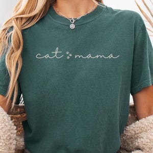 May include: A dark green short-sleeve t-shirt featuring the white cursive text "cat mama" with two small paw prints between the words. The shirt has a crew neckline and a relaxed fit, worn with light blue denim shorts and a beige textured cardigan.
