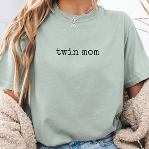 以下が含まれることがあります： 黒い文字で「twin mom」と書かれた、薄い緑色のTシャツ。デニムのショートパンツとクリーム色のカーディガンを合わせています。画像は柔らかく自然な雰囲気です。