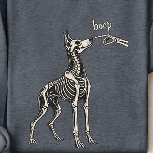 May include: A dark heather gray crewneck sweatshirt featuring a white graphic of a dog skeleton being touched on the nose by a skeletal finger. The word boop is printed in white above the dog's nose. The sweatshirt has long sleeves and a ribbed collar.