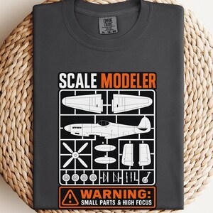 Può includere: T-shirt grigio scuro con grafica bianca e arancione. La grafica presenta un modello di aeroplano con la scritta "SCALE MODELER" e "WARNING: SMALL PARTS & HIGH FOCUS". La maglietta è esposta su una superficie circolare intrecciata.