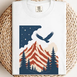 以下が含まれることがあります： 山とワシのデザインが施された白いTシャツ。赤と白のストライプの山、濃い青の松の木、星空が描かれています。Tシャツは、織られた円形の表面に折りたたまれています。