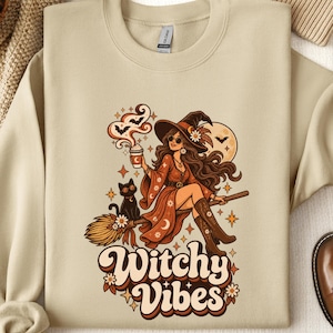 May include: A beige crewneck sweatshirt featuring a graphic print of a person wearing a witch hat and sunglasses, riding a broomstick with a black cat. The design includes a coffee cup, bats, stars, and the text Witchy Vibes in a retro font.